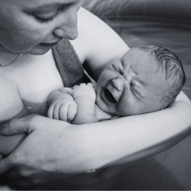 A mother holds a crying newborn in water, both looking at each other tenderly.